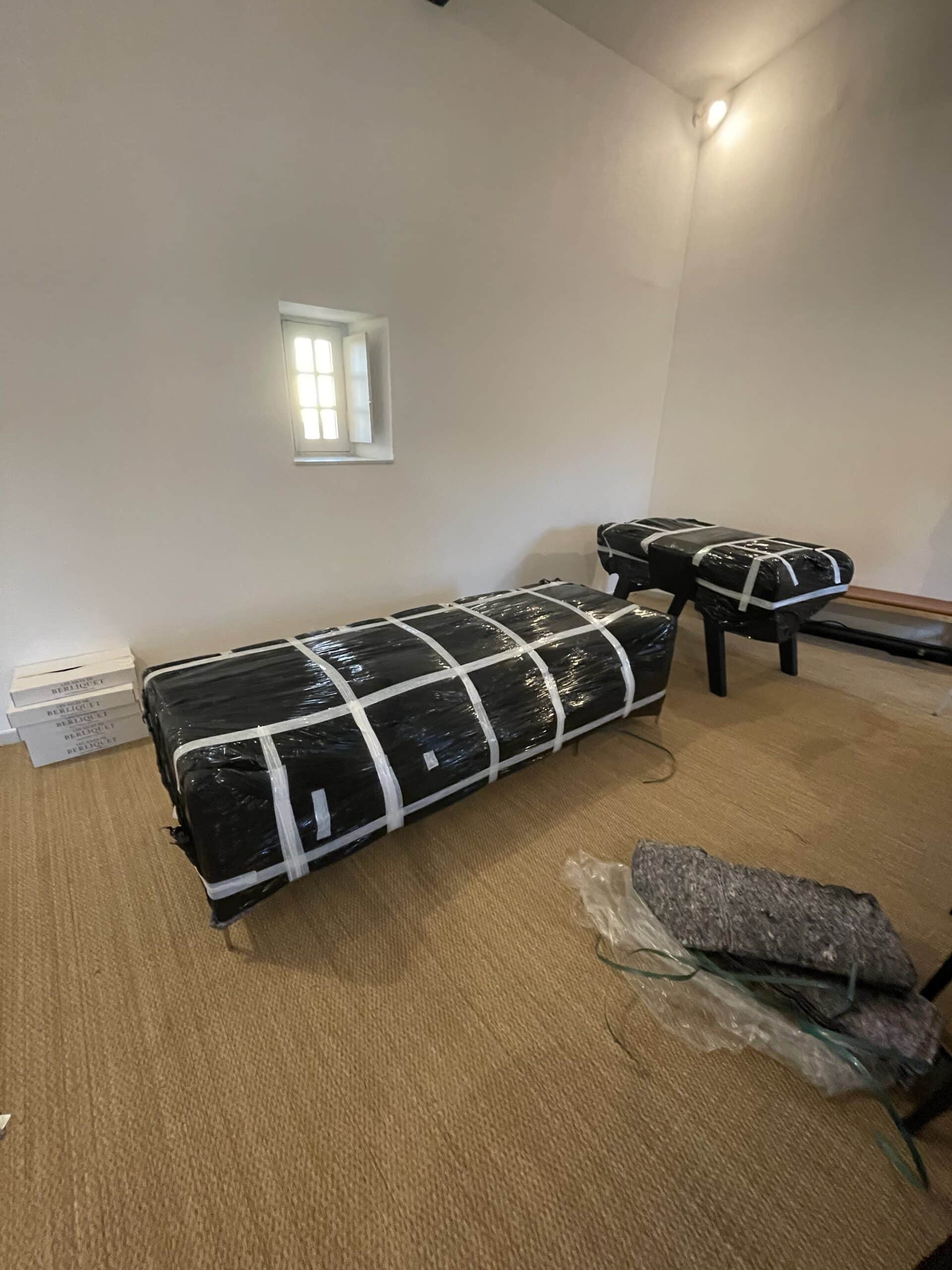 Deux gros articles emballés en plastique noir sur un tapis, près d'une fenêtre lumineuse et de boîtes "BERLIQUET".