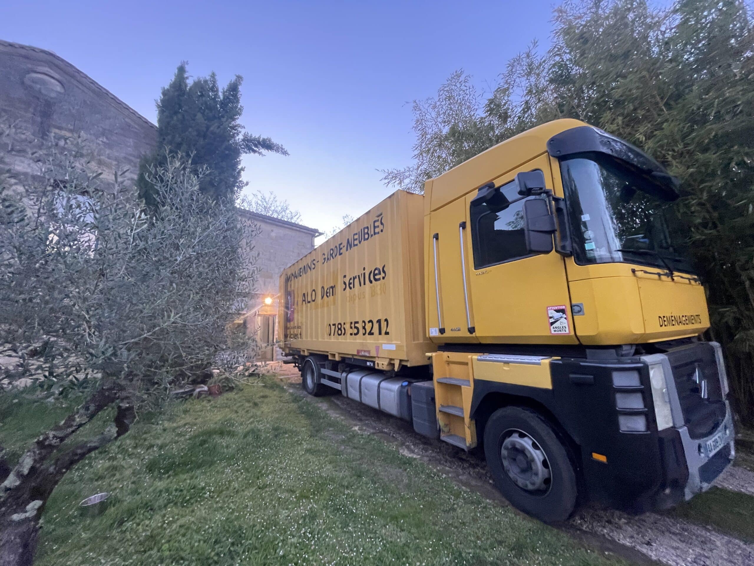 Camion jaune ALO Dem Services - Garde-Meubles 0785553212 stationné dans un jardin à la tombée du jour.
